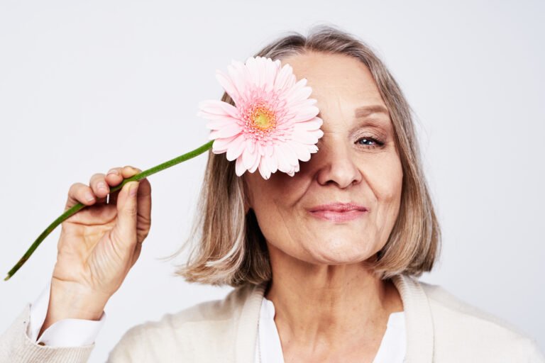 An older woman with grey bobbed hair smiles and holds a pink gerbera in front of one eye.